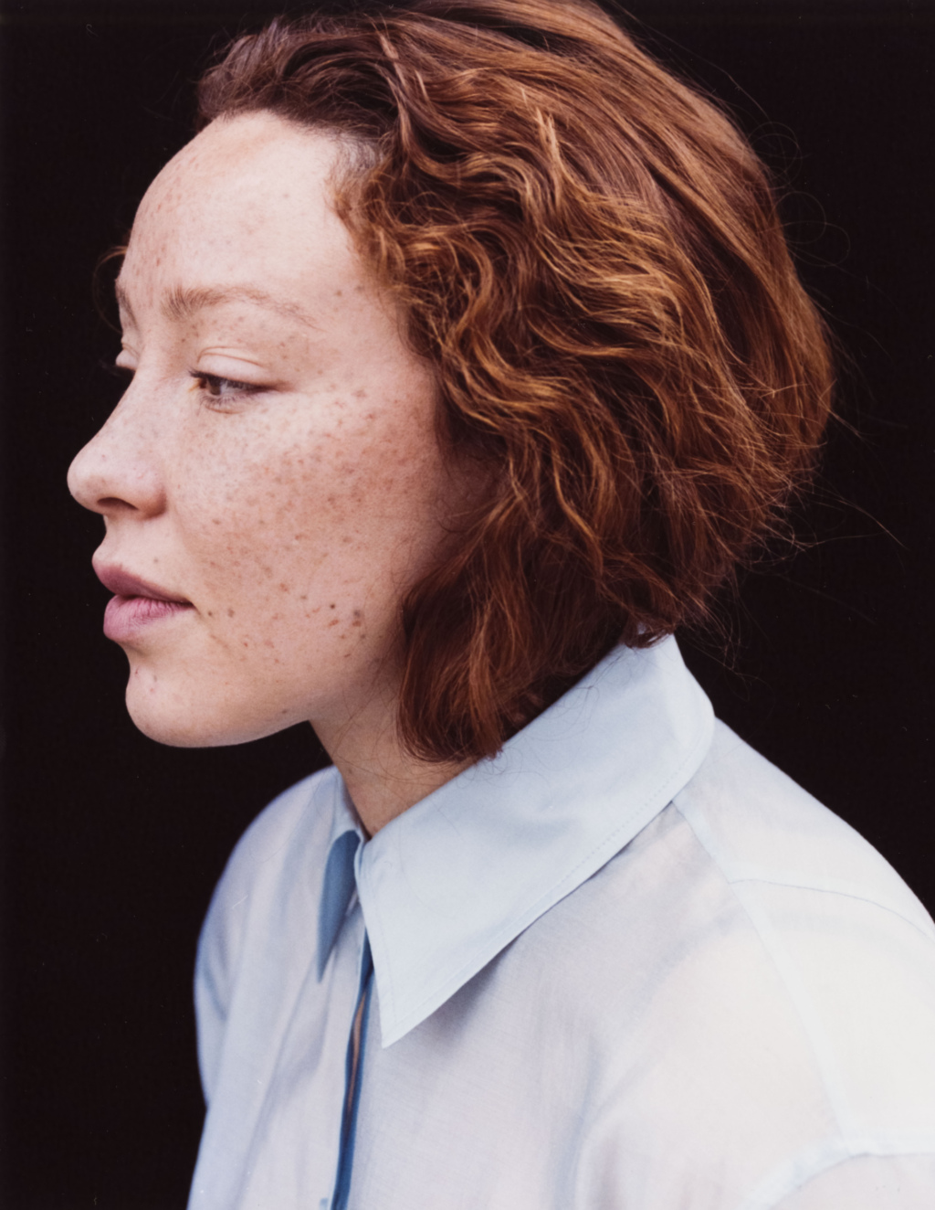 Portrait of young woman with red wavy hair facing left wearing a blue shirt